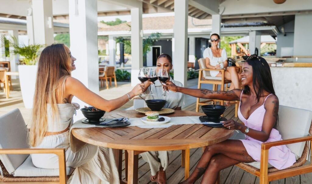 Women clinging half filled wine glasses, sitting around a round wooden table in a restaurant.