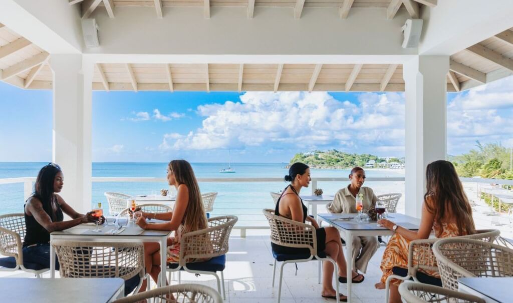 Guests dining at an open-air restaurant with white pillars and a high ceiling, enjoying a panoramic view of the sparkling blue ocean and distant land under a partly cloudy sky.