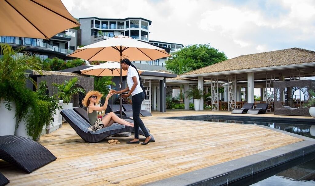 A woman relaxing on a sun lounger by a pool, being served a drink by a resort staff member - Tamarind Hills Resort and Villas - Antigua
