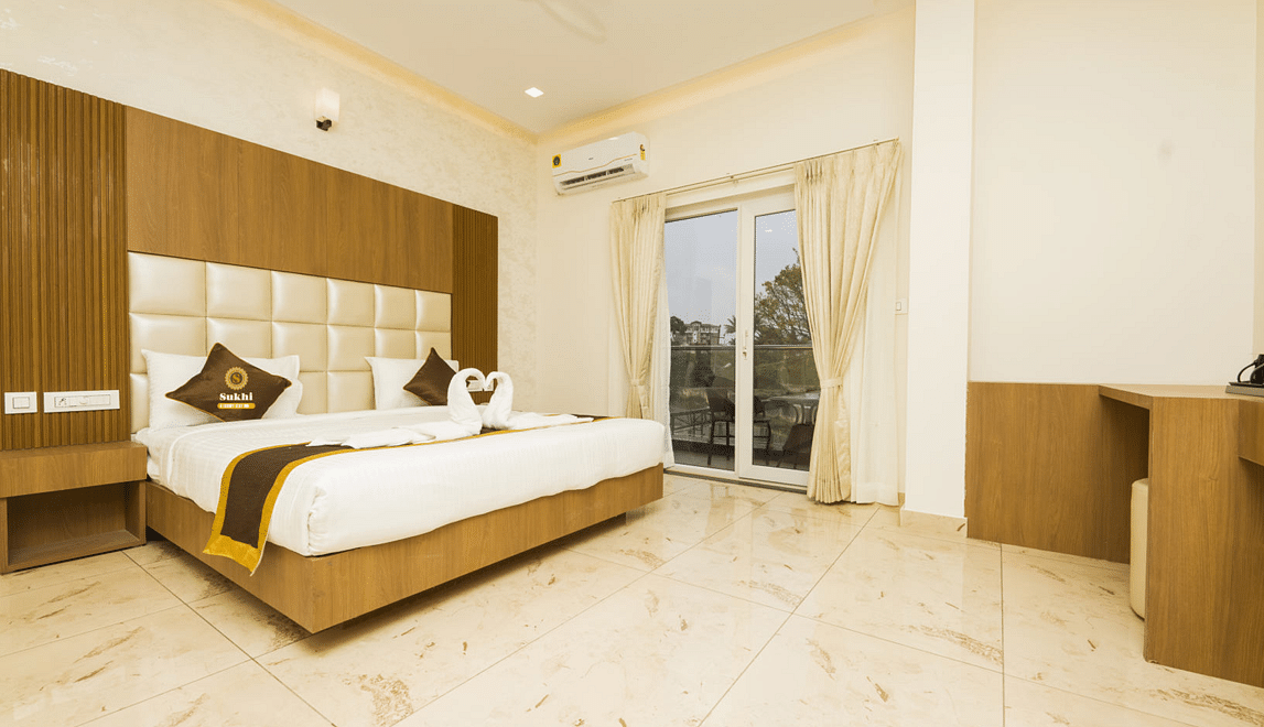 Super Deluxe Room With Balcony featuring a king size bed with a wooden headboard, and decorative pillows, alongside light-coloured walls, tiled flooring.- Sukhi - A Luxury Stay Inn, Chikkamagaluru