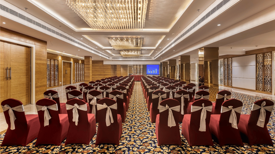 Spacious banquet hall with red chairs, round tables, and a chandelier-lit ceiling.