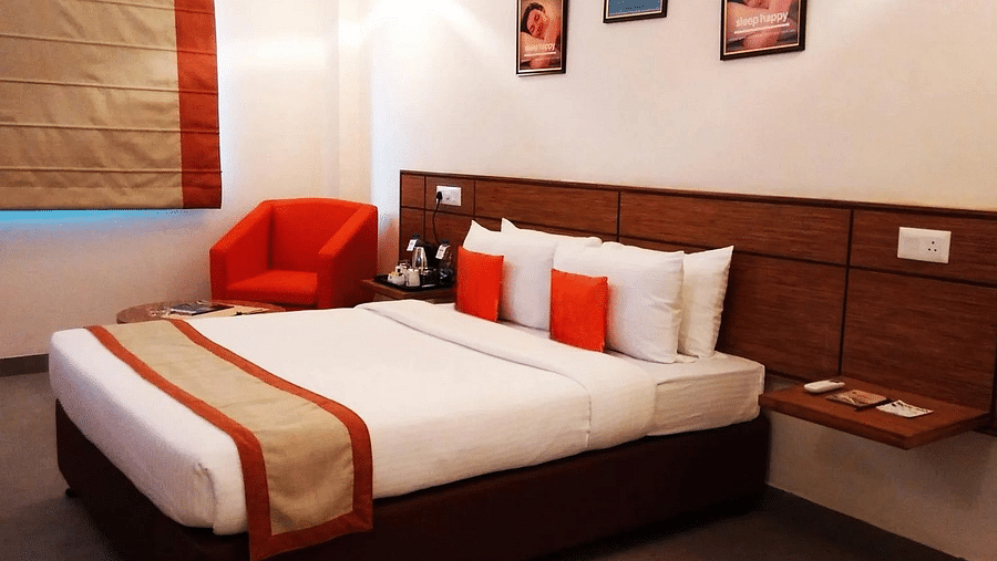 a close up shot of the queen size bed with a small red cushioned arm chair in the room - Max Hotel Jabalpur