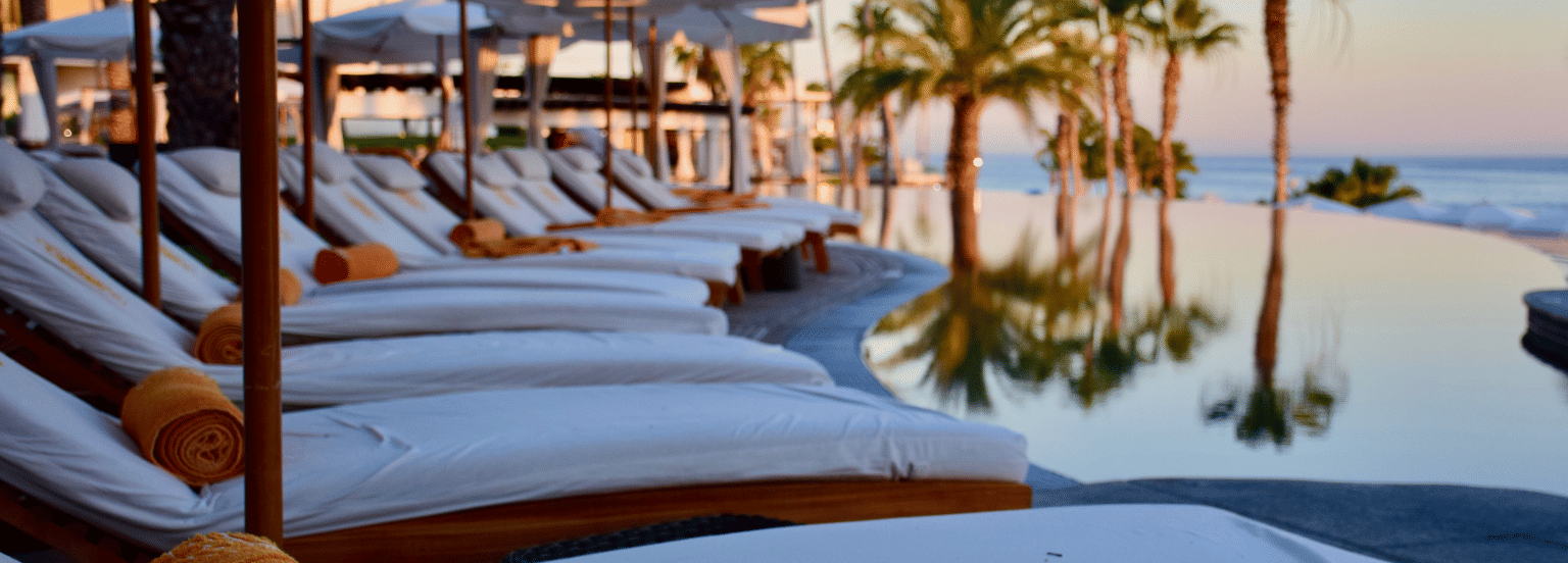 A serene waterfront view with luxurious poolside loungers overlooking a sunset-lit harbour, featuring yachts and palm trees.