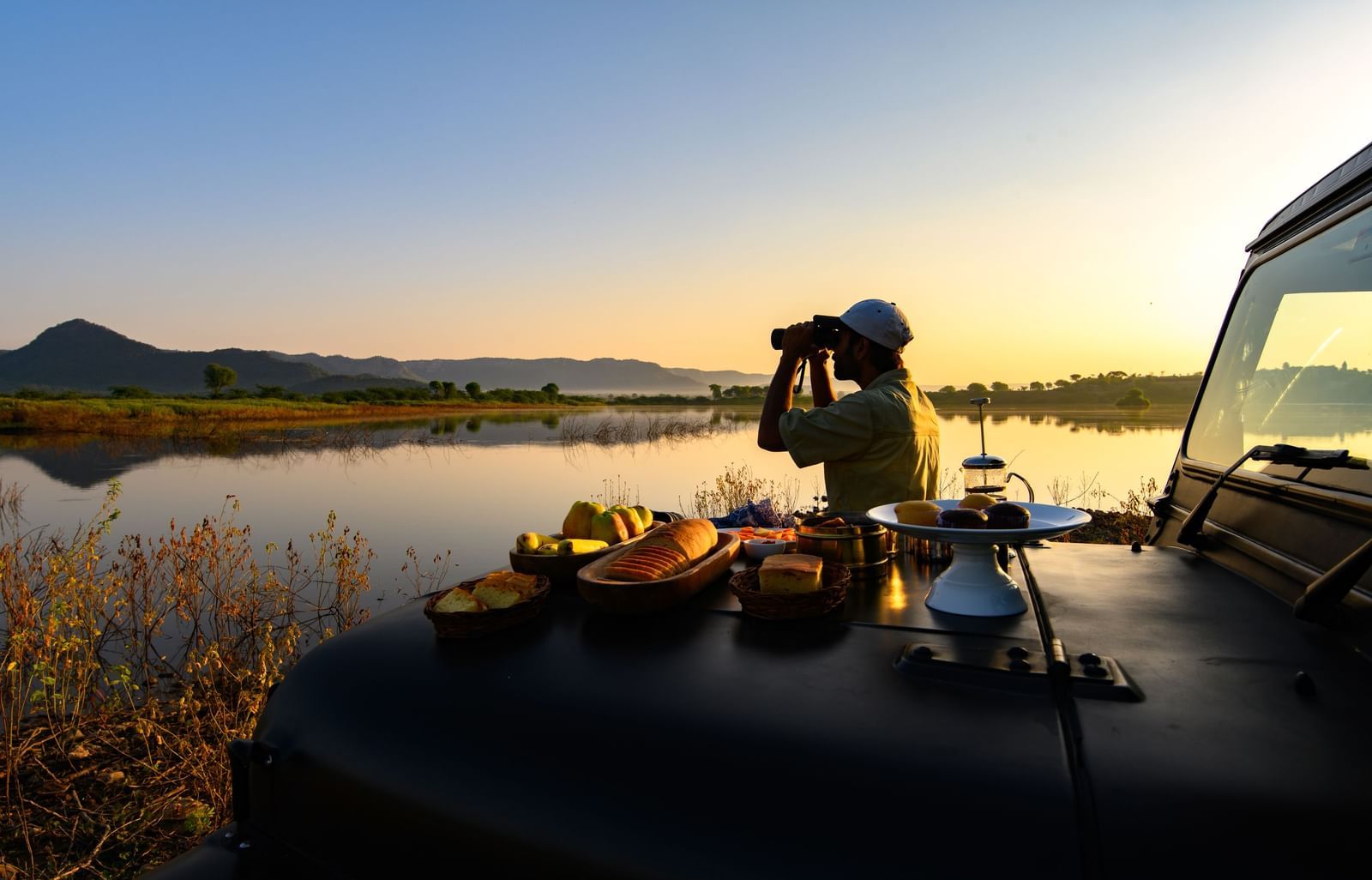 An image of a man standing near a lake with his jeep parked over looking the mountain - An image of a jeep parked amidst the rocky mountains - Utsav Camp Sariska