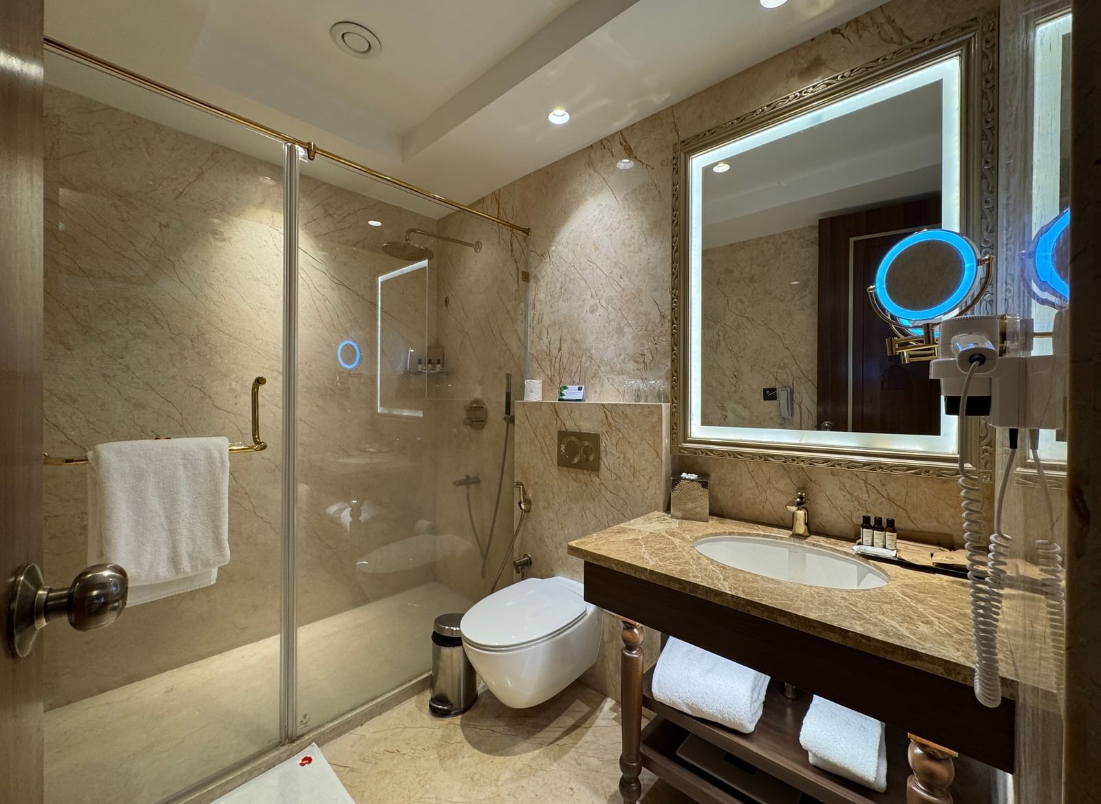 The washroom in the Deluxe Garden Twin room featuring a separate shower cubicle, a commode, a large sink, a back-lit mirror, and a magnifying shaving mirror - Kaldan Samudhra Palace