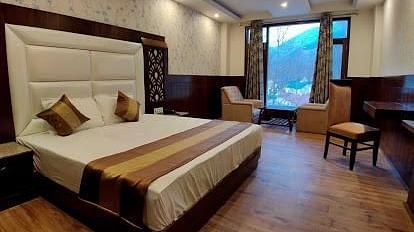 A spacious hotel room with a king size bed, two armchairs, a coffee table, a desk, and a window overlooking a garden | DLS La Serene Manali