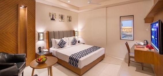A comfortable hotel bedroom featuring a double bed with white linens and patterned cushions, a wooden bedside table with a lamp and fruit bowl, a black armchair, a writing desk with a chair, and a television on the wall at Lords Inn Motikhavdi, Jamnagar.