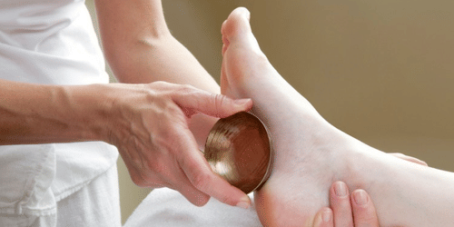 Therapist using a kansa bowl on a person’s foot during a massage session, moving the tool across the surface for treatment