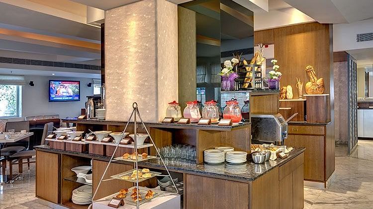 Buffet area at HHI Select, Bangalore featuring a variety of food displays, wooden counters, stacked plates, a TV screen, and modern interior decor. 