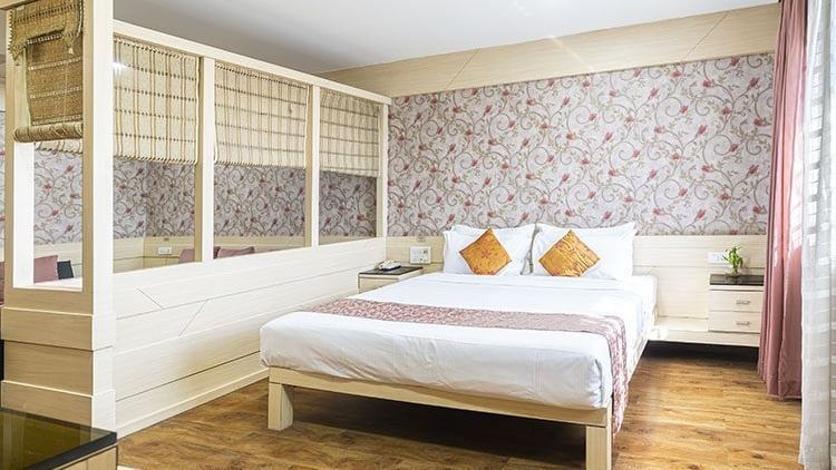 A well-lit suite featuring a king size bed with mustard cushions, floral wallpaper, wooden flooring, and a partial partition with glass panels and cream framing at Infiniti Hotel, Indore.