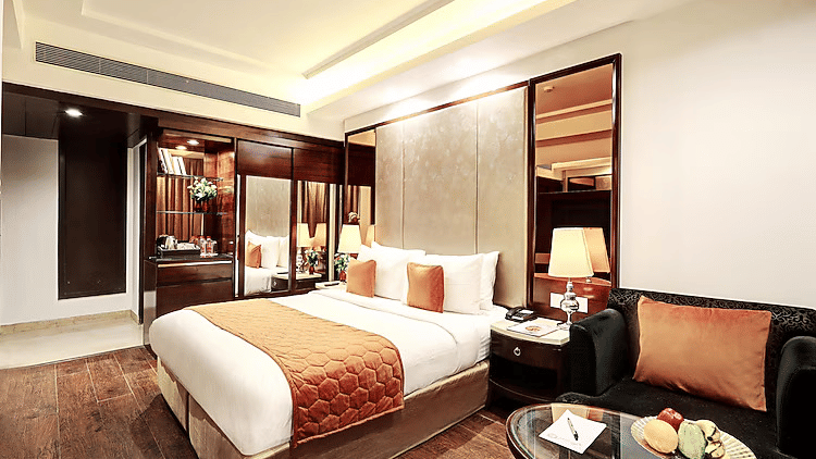 A well-lit boutique room showing a comfortable bed next to a black leather armchair and a glass coffee table with fresh fruit - Flute Boutique By Hotel 91, Jaipur