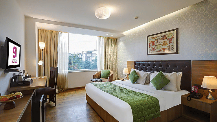 An inviting bedroom view showing a dedicated wooden workspace, a flat-screen TV, and a cozy armchair by the large window - Hotel 91 HUDA City Centre, Gurugram