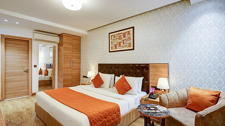 A stylish bedroom featuring a king size bed with an orange quilted runner, matching cushions, and a cosy corner armchair - Hotel 91 HUDA City Centre, Gurugram