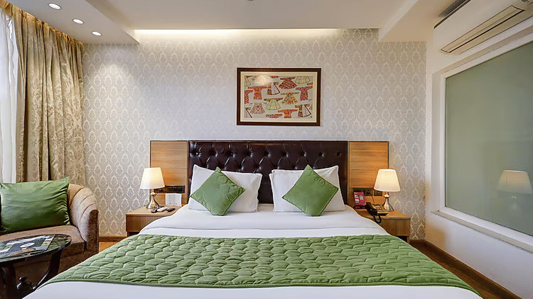 A front-facing view of a double bed with a dark tufted headboard, green quilted runner, and warm bedside lamp lighting - Hotel 91 HUDA City Centre, Gurugram
