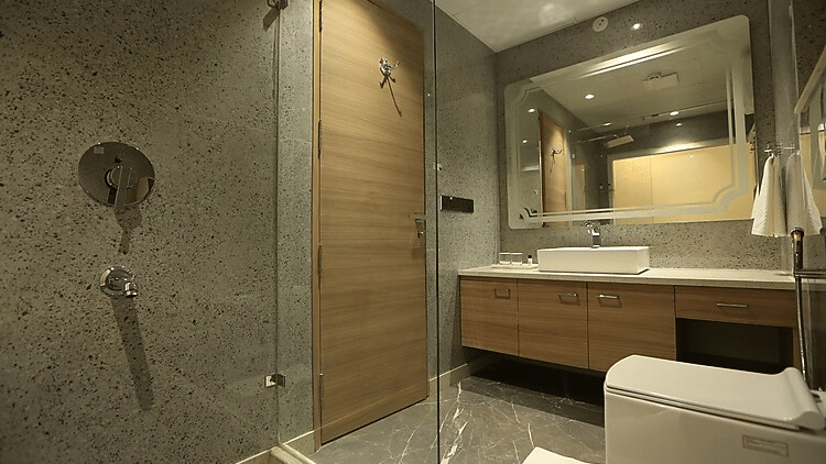 A sleek and clean bathroom with grey stone tiling, a large vanity mirror, a modern white sink, and a glass-enclosed shower area - Hotel 91 HUDA City Centre, Gurugram