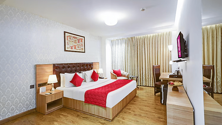 A wide-angle view of a premium guest room showing a bed with red pillows, light oak furniture, and elegant wall art - Hotel 91 HUDA City Centre, Gurugram