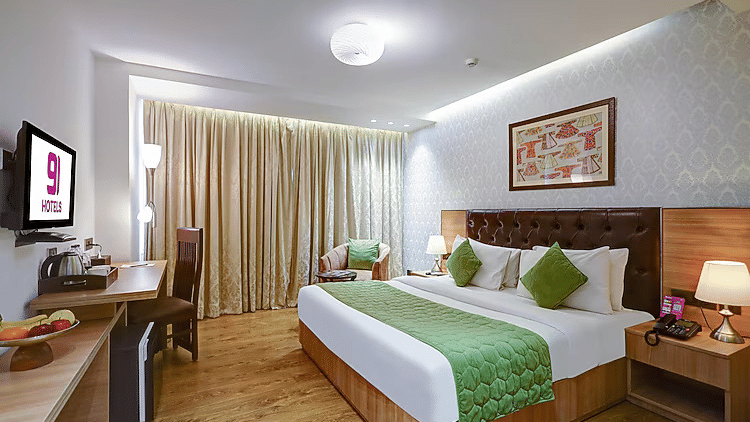 A spacious guest room with hardwood floors, floor-to-ceiling drapes, and a comfortable bed setup with green decorative pillows - Hotel 91 HUDA City Centre, Gurugram