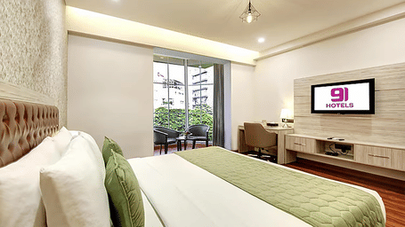 A large bed in a hotel room featuring a wall-mounted TV, a desk, and a door leading to a balcony | Hotel 91 Millennium HUDA, Gurugram