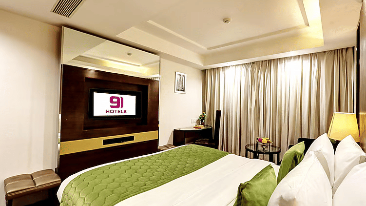 A clean and bright bedroom layout showcasing a wall-mounted TV with brand signage and a bed dressed in fresh white linens and green accents - Flute Boutique By Hotel 91, Jaipur