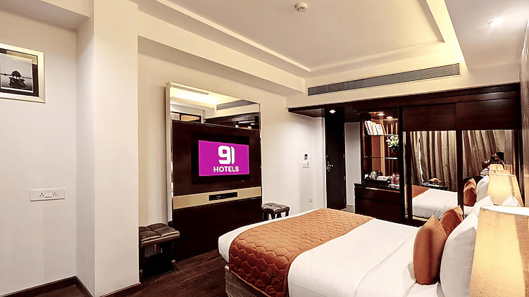 A bright guest room view focused on the wall-mounted flat-screen TV, minimalist white walls, and a cozy bed with orange accents - Flute Boutique By Hotel 91, Jaipur