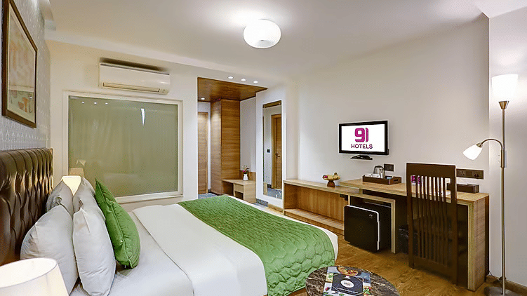 A brightly lit hotel room featuring a king size bed with a green quilted runner, matching cushions, and a wall-mounted TV over a wooden desk - Hotel 91 HUDA City Centre, Gurugram