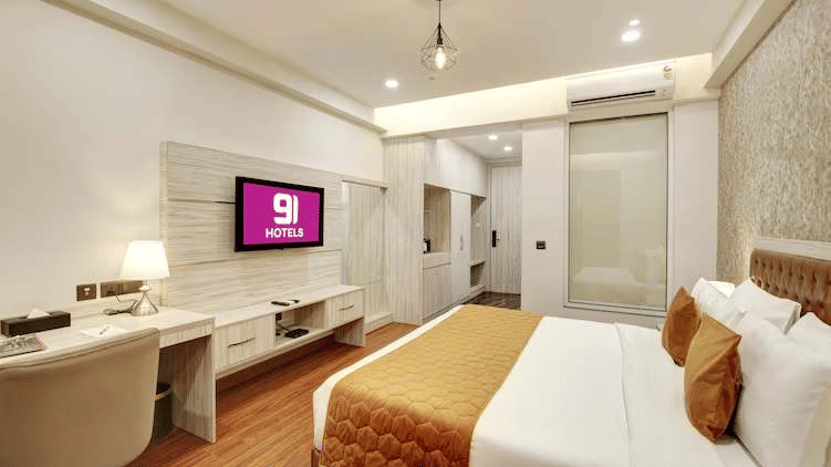 A hotel room featuring a large bed, a wall-mounted TV, a desk, and a bathroom entrance | Hotel 91 Millennium HUDA, Gurugram