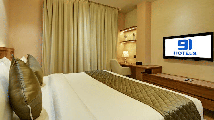 A wide-angle shot of the Deluxe Room at Hotel 91 Privé, Gurugram, featuring a neatly kept double bed with a wall-mounted television placed in front of it.