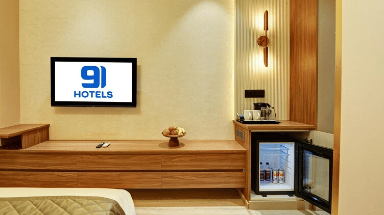 Front view of the wall-mounted television above the TV cabinet in the Deluxe Room at Hotel 91 Privé, Gurugram, with an open mini fridge beside it.
