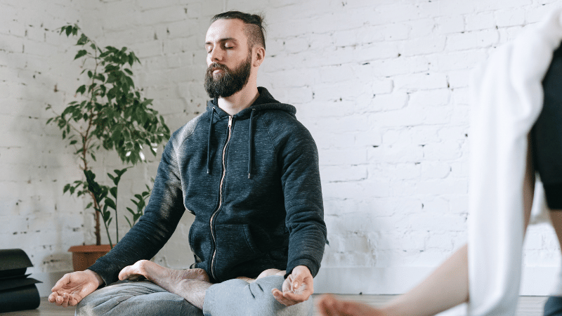 A person sitting cross-legged on a yoga mat with eyes closed, meditating in a calm indoor space with a plant and white brick wall in the background.
