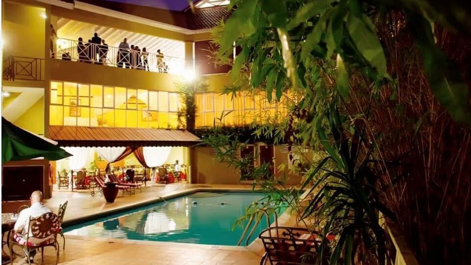An outdoor view of a hotel at night, featuring a swimming pool surrounded by palm trees and illuminated buildings | Altamont Court, Jamaica