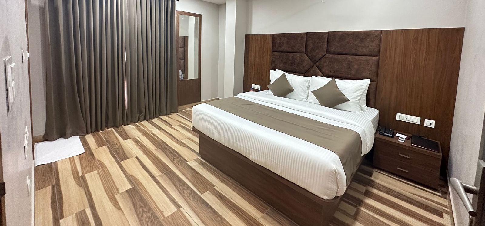 A room displays a double bed with white bedding, dark wooden headboard, and wooden flooring - Kankidham Lords Inn