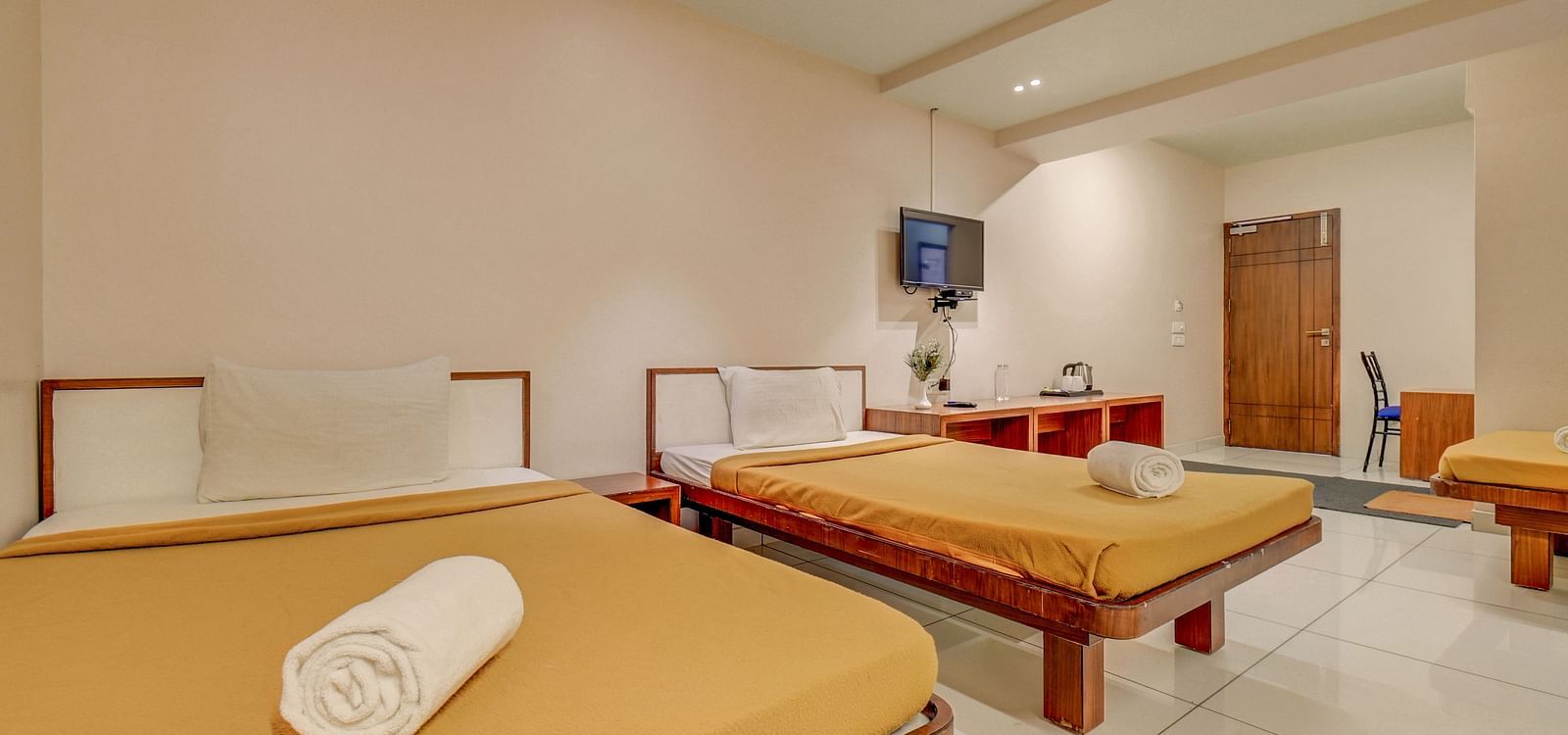 A spacious room at Hari Govinda’s JN Stay, Shakthinagar featuring neatly made single beds, a wall-mounted television, and a door at the furthest end