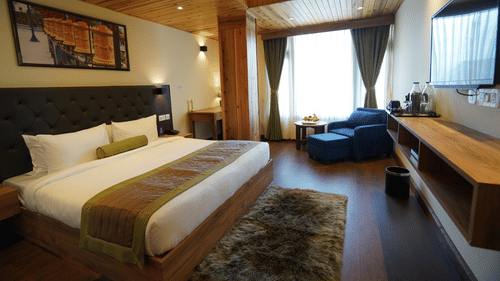 A luxurious room with a wood-panelled ceiling, a king bed with green and brown decor, a lounge chair by the window, and a wall-mounted TV - Mount Hotels