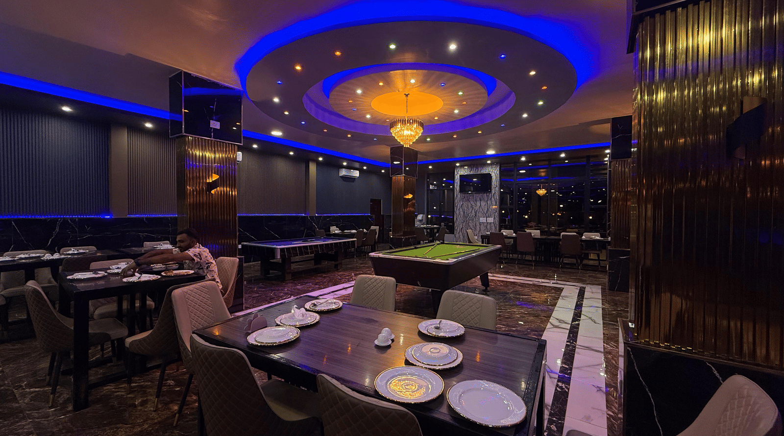 A dark, luxurious recreation room with a pool table, set tables, and illuminated by a central chandelier and blue neon ceiling lights at Luffu Club.