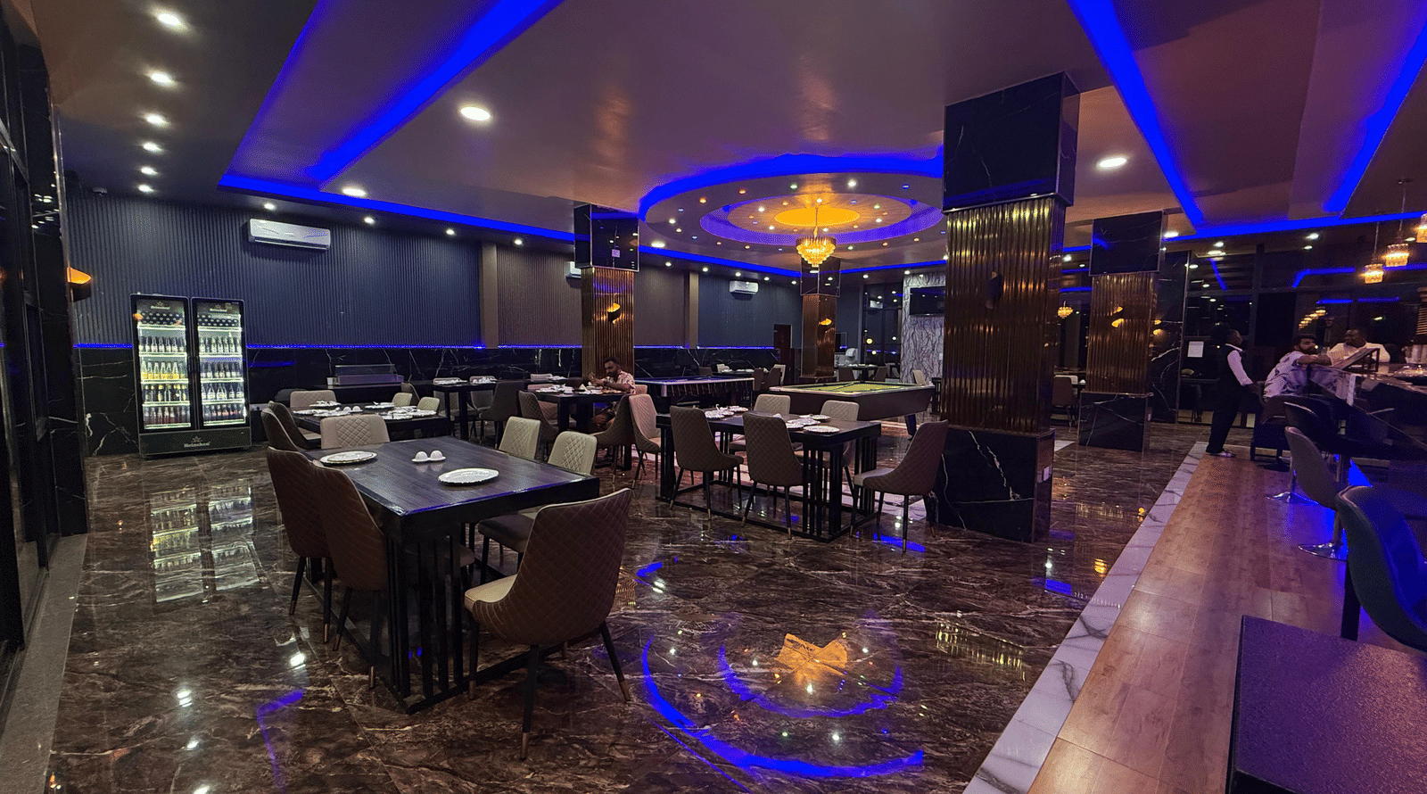 A panoramic view of a restaurant or bar interior, featuring dark decor, shimmering floors, and vibrant blue neon strip lighting at Luffu Club.