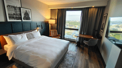 A high-rise guest room with a leather-panelled bed, white linen, a tan leather armchair, a desk beside a city-view window, and cityscape prints.