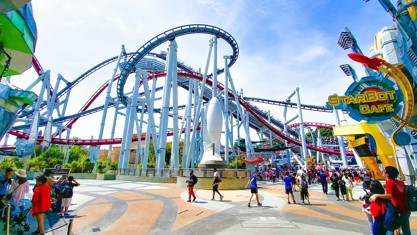 A wide-angle, low-angle shot of a roller coaster at a theme park on a bright, sunny day. 