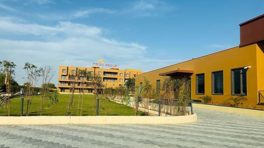 A spacious paved area and landscaped garden with buildings in the background at Pride Resort, Jodhpur under a clear blue sky.