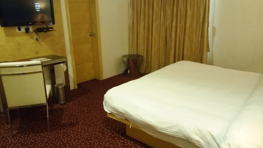a double bed room with a wall-mounted TV, a workstation with a chair, and a ceiling-to-floor windows covered in light yellow curtains