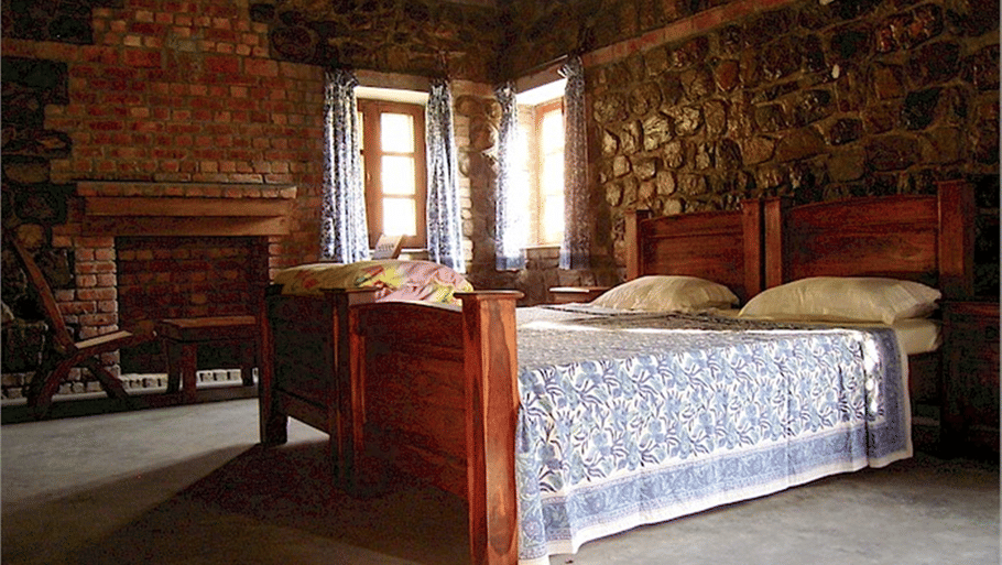 A rustic bedroom featuring a wooden bed frame, patterned blue coverlet, stone and brick walls, and sunlight filtering through windows.