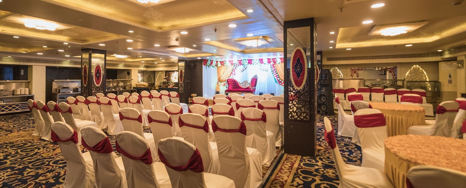 Maurya featuring rows of chairs with white covers and red sashes facing a stage area with draped fabric and lighting.