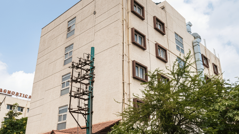 A clear angled view of the multi-storey VITS Select, Bengaluru building facade rising above dense surrounding trees under a bright cloudy sky, with a sign visible on the roof.