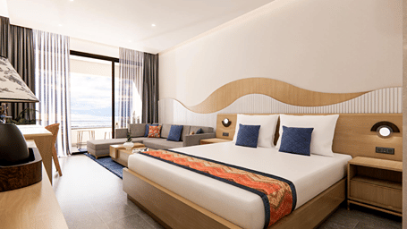Bright sea-facing hotel room with a king bed featuring orange and blue accents, a small sofa, and floor-to-ceiling glass doors opening onto a balcony.