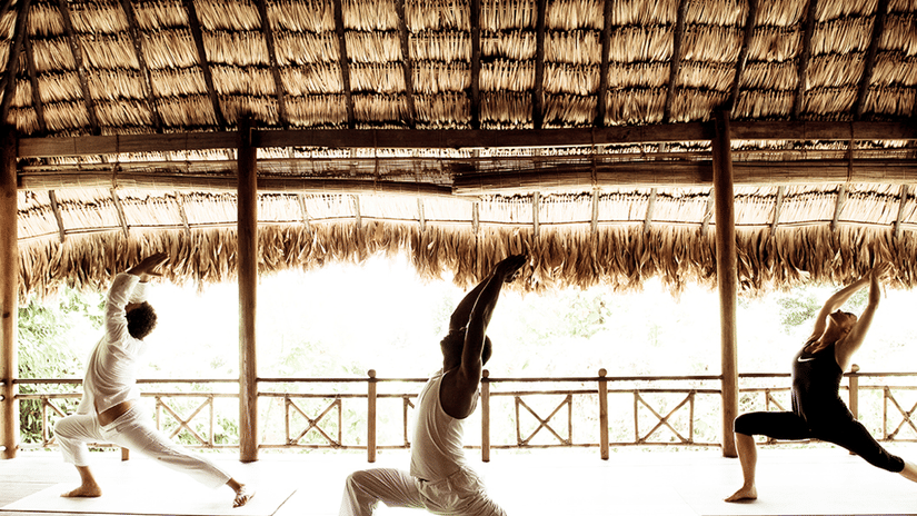 Three people are practicing yoga poses in an open-air studio with a thatched roof.