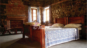 A spacious rustic bedroom with stone walls, wooden beds, and a brick fireplace in the corner near bright windows.