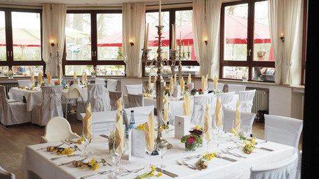 A dining room set up with multiple tables covered in linens and chair covers, featuring a central square table with a tall candelabra, cloth napkins, and windows in the background.