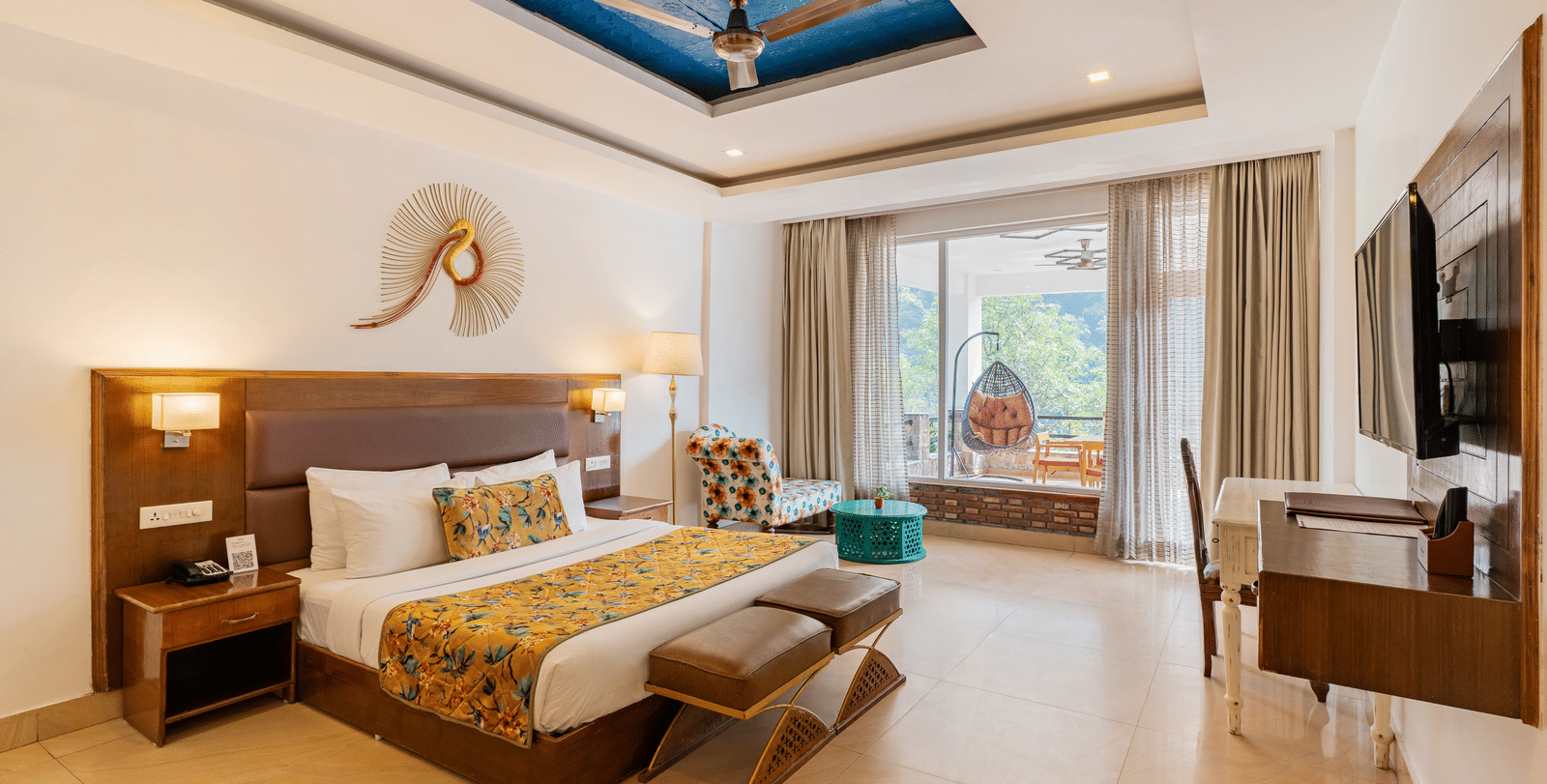 A well-lit bedroom featuring a big bed facing a television screen, with an attached balcony featuring a standing swing chair overlooking the trees outside | Summit by the Ganges Beach Resort & Spa, Rishikesh