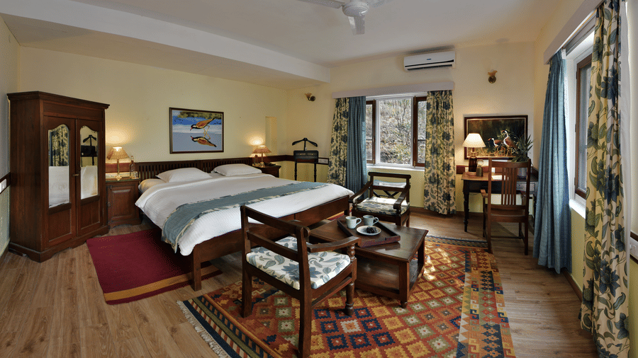 Refined bedroom with polished wooden flooring large bed seating corner and ample daylight ensuring a serene stay at Neemrana’s Glasshouse on The Ganges with a balance of comfort and elegance