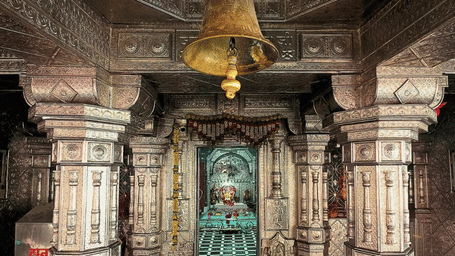 A hall inside a temple. A bell hangs from the ceiling in the centre. Pillars stand along the sides of the hall. A statue sits in a shrine in the background. A person sits on the floor near a pillar.