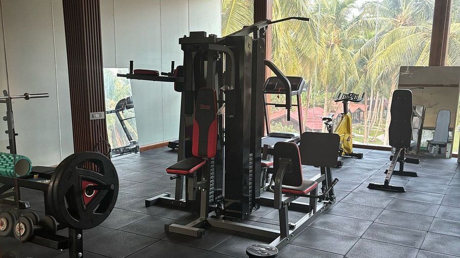 A gym with various strength training equipment, including a multi-functional weight machine and barbells, set in a spacious room with large windows overlooking palm trees and a red-roofed structure - Silver Sand Beach Resort, Havelock.